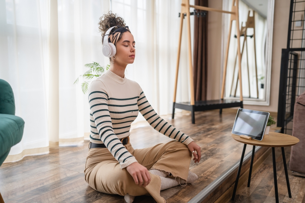 How to Avoid Time-Wasting Activities in Your Remote Workday: Picture of a female millennial using headphones for online guided meditation practicing mindfulness yoga with eyes closed on the floor at home real people self care concept.