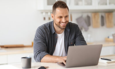 Secrets to Staying Healthy While Working Remotely: Young happy man independent contractor working on laptop and smiling, enjoying remote work, sitting at home kitchen interior, free space