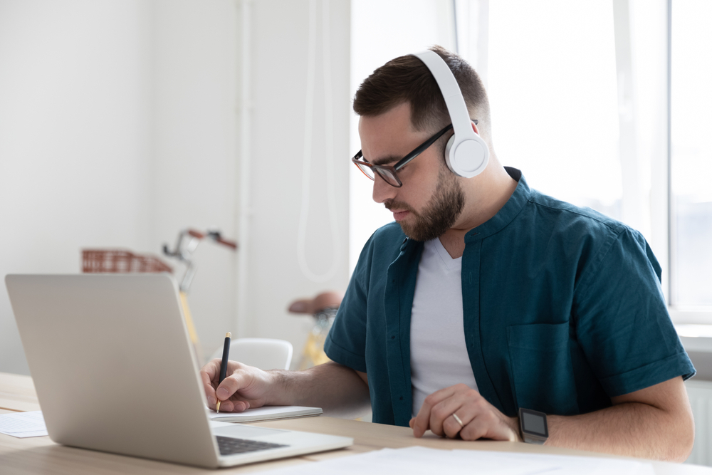 Things to Know Before You Start a Remote Job: A picture of a focused young freelancer in glasses wearing wireless headphones, watching educational webinar lecture seminars on a laptop online, and writing down notes in a modern home office. 