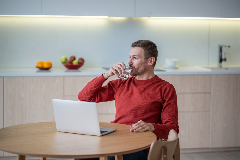 Secrets to Staying Healthy While Working Remotely: Picture of a young man drinking water while working remotely.