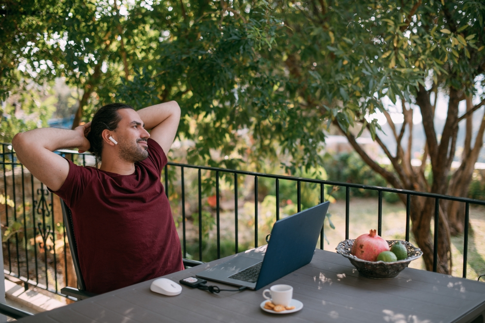 Things to Avoid When Working from Home: A young, handsome man is sitting relaxed at a laptop at a table on a veranda in a green garden. 