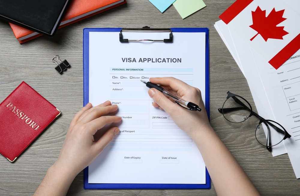 Canada Visa Rejection Triggers to Avoid at All Costs: Woman filling visa application form to Canada at wooden table, top view 