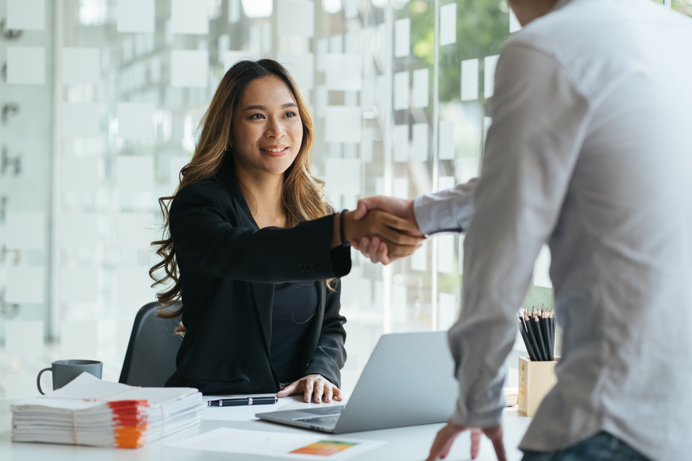 Self-Employment vs Employment in the UK: Picture of a smiling asian manager sitting at her desk in an office shaking hands with a job applicant after an interview 