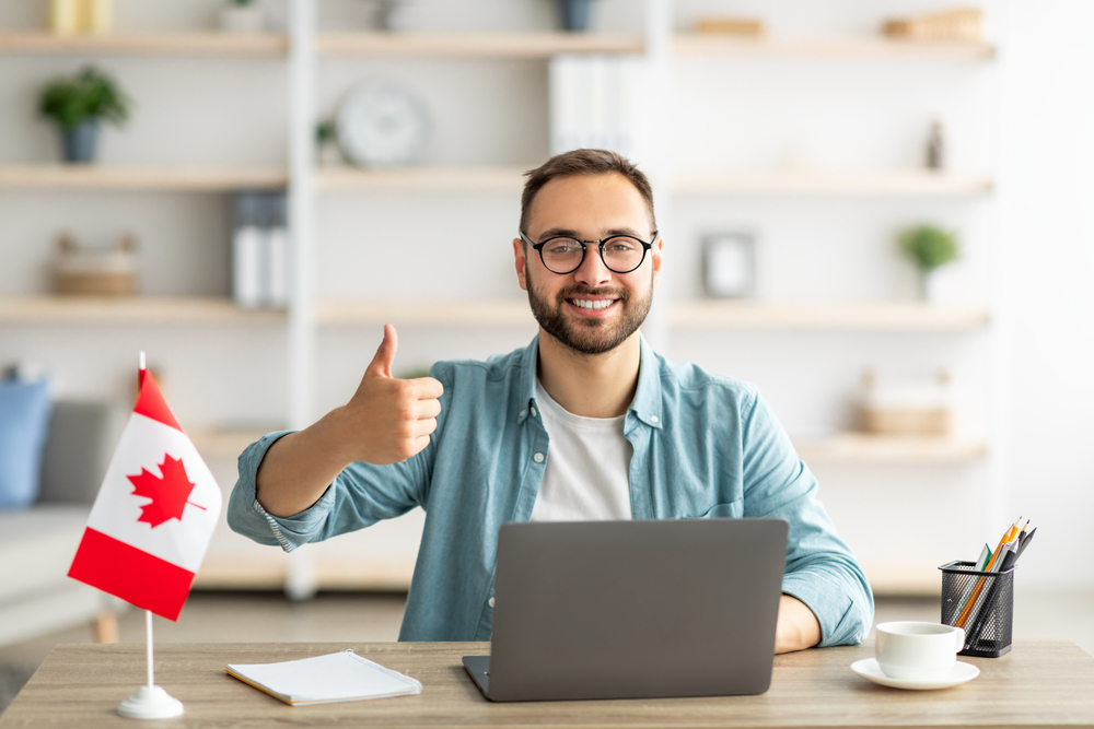 Canada Post-Graduation Work Permit vs Skilled Worker Visa: Happy young guy sitting at table with flag of Canada, using laptop computer, showing thumb up gesture at home.