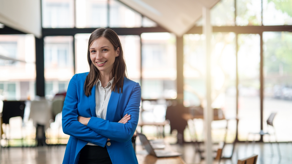 Associate Bankers Job:A professional woman with a confident smile stands in a bright, airy office space, dressed in a smart blue blazer with arms crossed. 