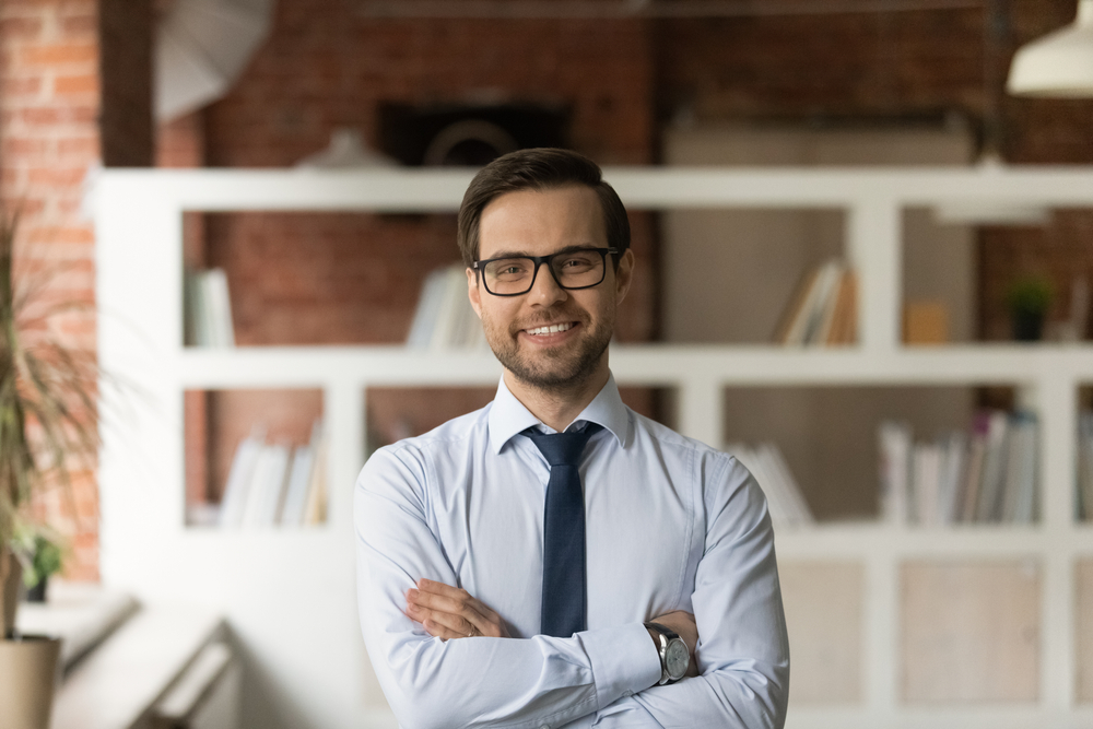 How salary negotiation in Canada works: Portrait of sincere smiling confident young 30s male CEO executive manager in eyeglasses and formal wear posing in modern office room