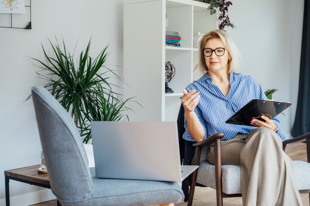 Top 10 Fully Remote, In-Demand Jobs That Pay Over $100K: Professional Psychotherapy online. Middle aged female Psychologist having video call consultation on laptop while sitting on armchair In office. Specialist talking, counseling.